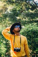 Young Latin American man in a forest wears a yellow sweater with a camera and a hat, looking around. travel and natural recreation concept. photo