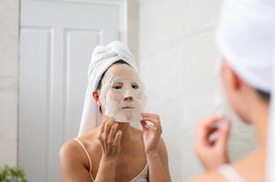 An Asian woman is applying a facial mask pad to her face in front of the bathroom mirror. photo