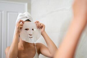 An Asian woman is applying a facial mask pad to her face in front of the bathroom mirror. photo