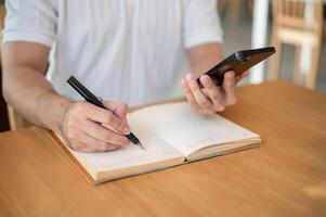 A cropped shot of an Asian man holding his smartphone while taking notes in his notebook. photo