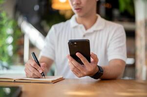 A cropped shot of an Asian man holding his smartphone while taking notes in his notebook. photo