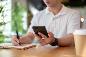 A cropped shot of an Asian man holding his smartphone while taking notes in his notebook. photo