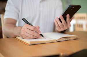 A cropped shot of an Asian man holding his smartphone while taking notes in his notebook. photo