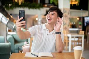 A cheerful Asian man is enjoying a call with his friend on his smartphone in a coffee shop. photo