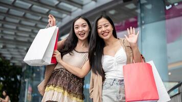 Two cheerful Asian women stand in front of the mall, carrying their shopping bags and smiling. photo
