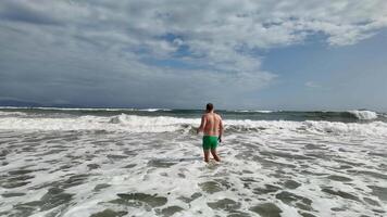A Caucasian man in green swim trunks stands in ocean waves under a cloudy sky, evoking themes of summer vacation and relaxation video