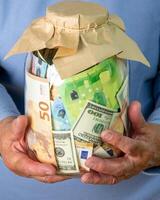 Close-up image of a persons hands holding a glass jar filled with various Euro and US dollar bills photo