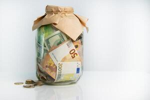 A glass jar filled with US dollar and Euro bills with a few coins scattered around it photo