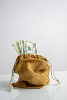 A close-up of a brown burlap sack with a drawstring top, filled with US dollar bills. The sack is sitting on a plain white surface photo