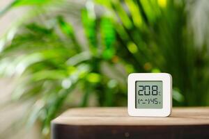 White Digital Thermometer Displaying Temperature and Humidity on Wooden Table With Green Plant in Background photo