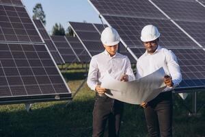 Two Engineers Wearing Hard Hats Analyzing Blueprints at a Solar Panel Field on a Sunny Day photo