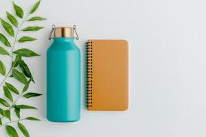 Eco-friendly flat lay featuring a teal reusable bottle, amber notebook, and green leaves on a neutral background photo