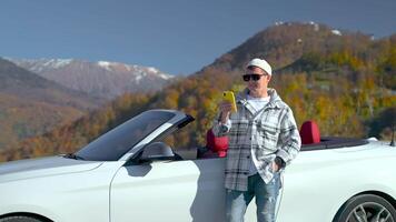 joyful traveler standing near his luxury auto cabriolet in high mountain area, using smartphone video