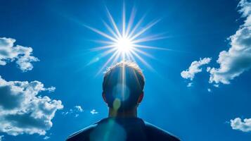 A striking silhouette of an individual uniquely stands against the backdrop of a bright sun, with its golden rays spilling over a clear blue sky, while fluffy white clouds drift gently by photo