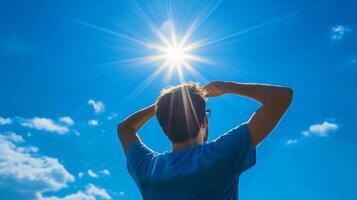 A person in a blue shirt stands outside on a lovely day, shielding their eyes from bright sunlight under a clear sky with fluffy clouds, enjoying this moment in nature, feeling free and happy photo