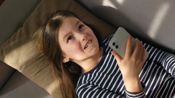 caucásico europeo pequeño niña acostado en sofá participación utilizando móvil teléfono teléfono inteligente niño artilugio en línea mensajes de texto SMS social redes sonriente riendo disfrute acecho gracioso Internet chateando niño foto