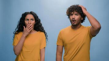 Multiracial shocked scared afraid couple amazed shocked Hispanic woman hiding behind Indian man astonishment wonder scare terrible horror ugly reaction blue studio background shock astonish fright photo