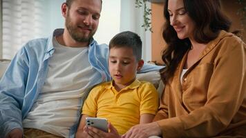 Happy caucasian family dad mom kid son relax on sofa together looking at smartphone screen using funny mobile application for play on phone having fun with technology watch on Internet at home photo