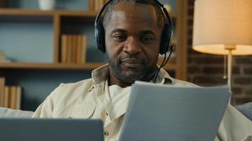 African american man teacher businessman freelancer in headset checking documents entering data in laptop remote working at home using computer application paying bills processing online information photo