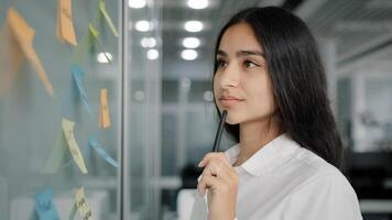 Arabian business lady managing daily tasks stand near board in office write reminder information instruction on paper sticky notes read stickers create to-do list analyze work organize corporate goals photo