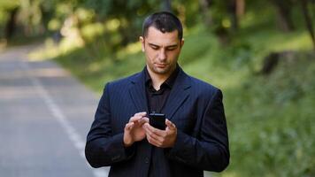 Serious adult caucasian man user stand outdoors in park wears black formal suit hold mobile telephone scrolling slide pages on smartphone screen online information texting message social business chat photo