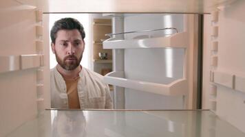 Hungry Caucasian bachelor man looking for food in empty fridge finding no meal products feeling sad disappointed poor unemployment need delivery service POV point of view from inside refrigerator photo