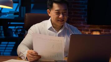 Middle-aged happy smiling businessman working with laptop and documents at dark night office Asian Korean man financial analyst accounting business using computer app at nighttime insomnia deadline photo
