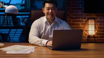 Middle-aged happy smiling businessman working with laptop and documents at dark night office Asian Korean man financial analyst accounting business using computer app at nighttime insomnia deadline photo