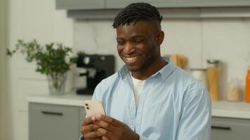 contento sonriente africano americano hombre jugando con móvil teléfono a hogar cocina sonrisa alegre positivo riendo birracial masculino étnico chico usuario comunicado con amigos social medios de comunicación red teléfono inteligente aplicación foto