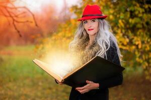 Halloween a woman with a book of magic spells in a witch's hat is conjuring, performing witchcraft against the background of an autumn forest. photo