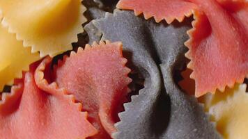 Uncooked Vibrant Colored Farfalle Pasta. A Culinary Canvas of Multicolored Bow-Tie Macaroni, Textured Background - Top View, Angular Rotation. Turning Colored Dry Pasta. Raw Macaroni - Rotating video