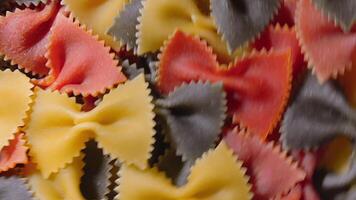Uncooked Vibrant Colored Farfalle Pasta. A Culinary Canvas of Multicolored Bow-Tie Macaroni, Textured Background - Top View, Angular Rotation. Turning Colored Dry Pasta. Raw Macaroni - Rotating video
