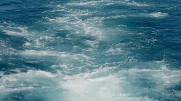 Close-up of waves and splashes with white sea foam forming patterns on surface of deep blue water. Ship trails in blue ocean video
