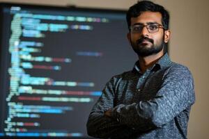 Indian programmer standing with crossed arms in front of computer screen displaying code photo
