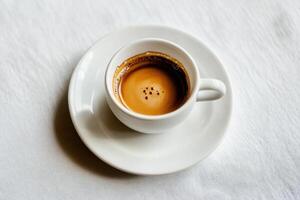 Small white cup of coffee resting on a saucer on white tablecloth photo