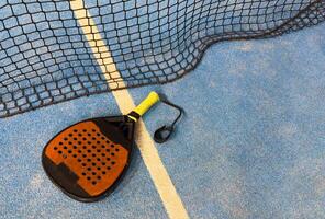 A padel or paddle tennis racket and a bright yellow tennis ball lie on a blue court. The net and a blurred figure of a player are visible in the background. photo