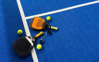 A padel or paddle tennis racket and a bright yellow tennis ball lie on a blue court. The net and a blurred figure of a player are visible in the background. photo