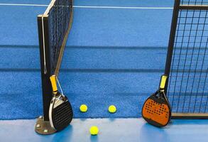 A padel or paddle tennis racket and a bright yellow tennis ball lie on a blue court. The net and a blurred figure of a player are visible in the background. photo