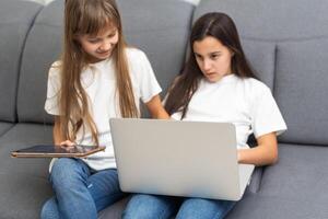 Cute little girl and older sister playing together smiling and having bonding time using a laptop on couch at home. Happy family Siblings relationship and digital technology lifestyle concept. photo
