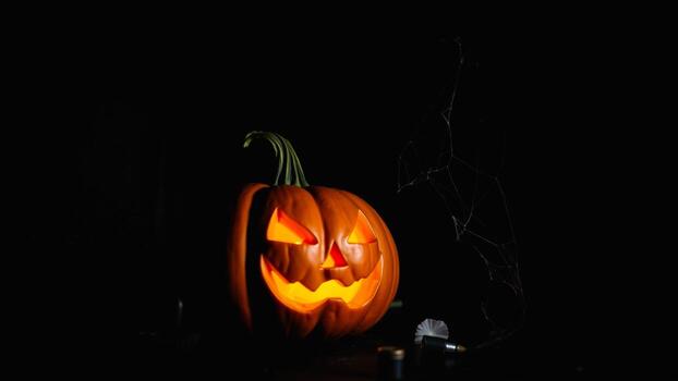 lit jack-o'-lantern with a glowing smile sits on a dark surface with a cobweb photo