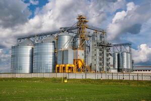 large grain elevator with multiple cylindrical metal silos, structural elements, and a complex system of pipes. It stands against a blue sky, with a field in the foreground after harvest photo