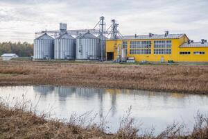 large grain elevator with multiple cylindrical metal silos, structural elements, and a complex system of pipes. It stands against a blue sky, with a field in the foreground after harvest photo