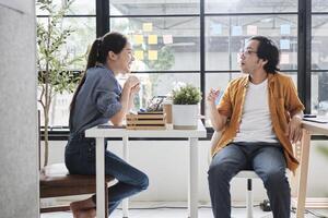 Two Asian workers sitting in chairs at working desks, collaborating and talking at daylight windows in casual small business office, online business job coworkers, positive relationship colleagues. photo