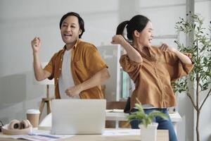 Happy working in casual small business office. Two Asian startup company colleagues enjoy dancing and singing songs together, relaxing from freelance jobs, humor, and funny coworker team in workplace. photo