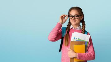Time to learn. Confident smiling teenage girl adjusts her glasses and holds notebooks and charts isolated on blue background, studio shot photo