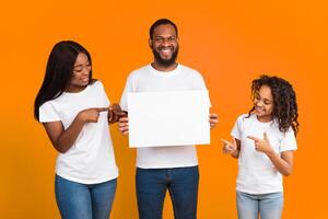 Place For Your Ad. Portrait of smiling black man holding empty blank board, family pointing fingers at square paper for mockup template, free space for text or design isolated on yellow studio wall photo