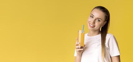 Portrait of smiling young woman with glass of orange vitamin juice in hand, holding glass with straw, posing over yellow background, free space photo