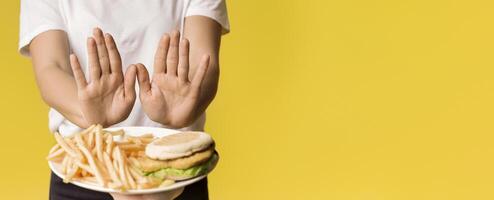 Unrecognizable Woman Rejecting Consume Plate With Junk Food, Showing Stop Gesture With Open Palms, Yellow Background, Cropped Image photo