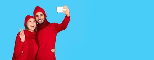 Happy couple in winter hats and scarfs taking selfie on smartphone, joyful young man and woman capturing photos together on mobile phone, standing isolated over blue studio background, copy space