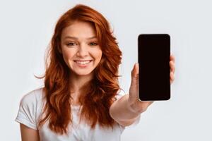 Mobile App. Smiling Ginger-Haired Girl Showing Cellphone Blank Screen Recommending Application On White Background. Mockup, Studio Shot photo